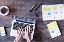 Hände tippen auf Laptop, daneben Smartphone, Tablet und Skizzen auf Holz­tisch