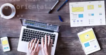 Hände tippen auf Laptop, daneben Smartphone, Tablet und Skizzen auf Holz­tisch