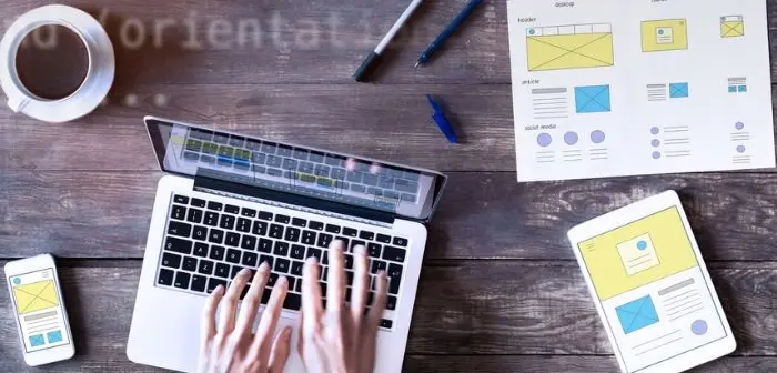 Hände tippen auf Laptop, daneben Smartphone, Tablet und Skizzen auf Holz­tisch